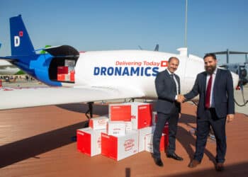 Two men shake hands in front of a cargo drone with parcels stacked next to them.