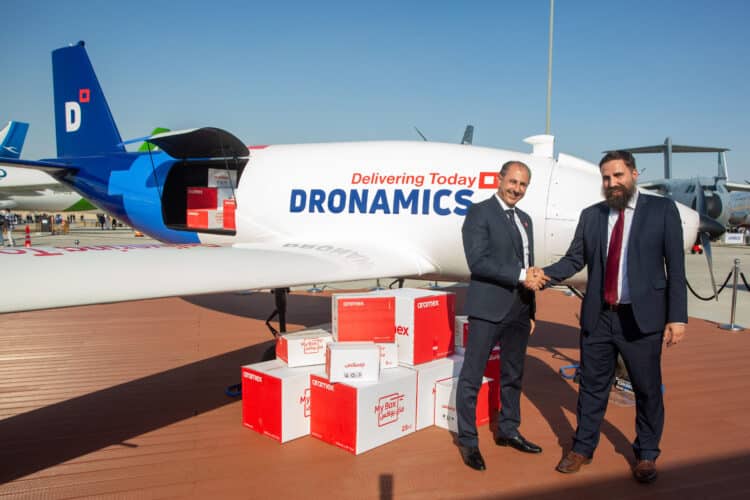 Two men shake hands in front of a cargo drone with parcels stacked next to them.