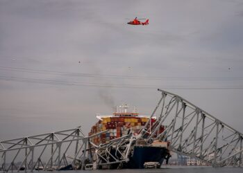 A helicopter flies over a bridge that has collapsed onto a container vessel