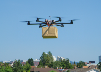 A drone carrying a large brown package flies over rooftops