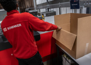 A man wearing a DB Schenker jacket stands alongside an empty box in a warehouse