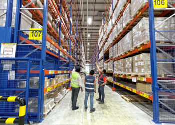 Three men stand in a warehouse, pointing to the top shelf of a cargo rack
